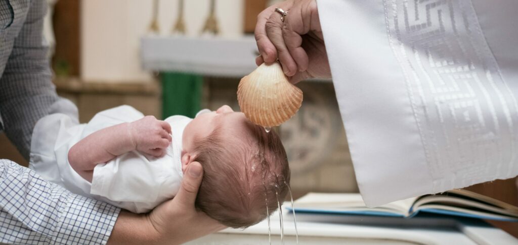 Baby wird, von Vater gehalten, durch einen Geistlichen über einem Becken mit Wasser beträufelt.