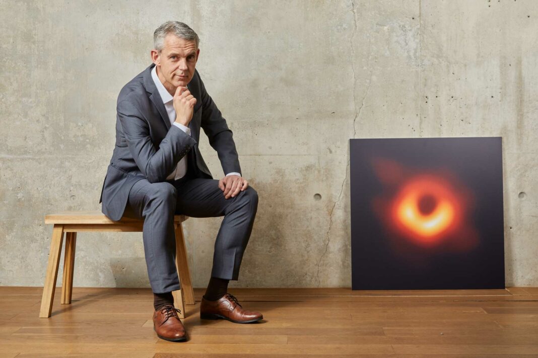 Der Physiker Heino Falcke sitzt neben dem Bild eines Schwarzen Lochs.