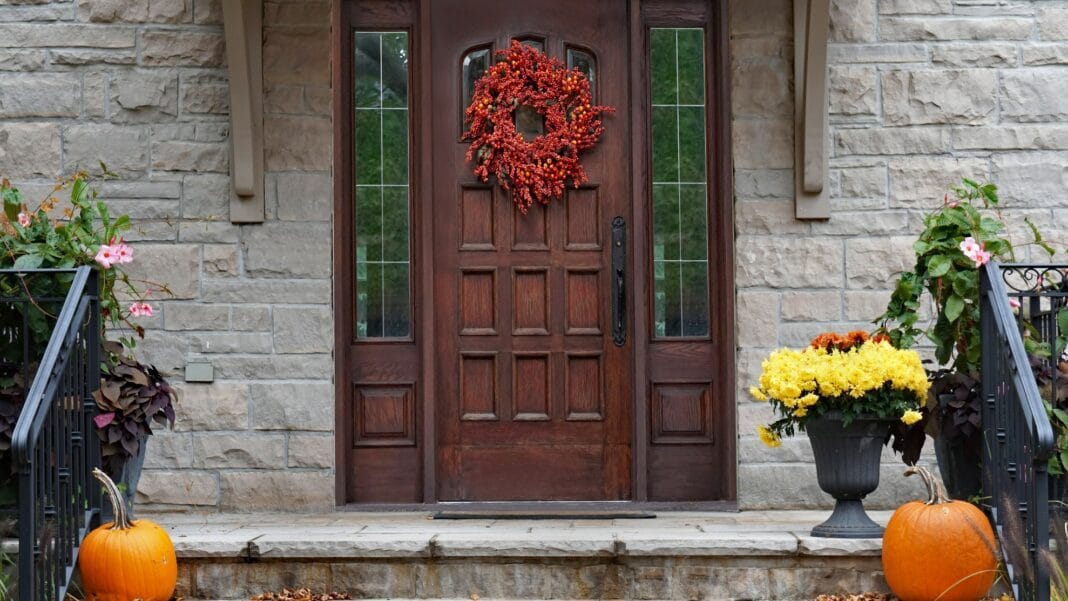 Eine herbstliche dekorierte braune Holzhaustür. Auf den Treppenstufen stehen Blumen und Kürbisse.