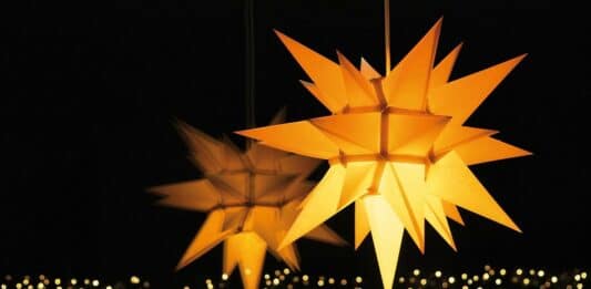 Ein gelb leuchtender Herrnhuter Stern, der sich in einem Fenster spiegelt. Unter ihm sieht man eine Lichterkette.