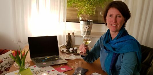 Eine Frau sitzt an einem dekorierten Tisch und hebt ein Weinglas in die Höhe. Auf dem Tisch steht ein Laptop.