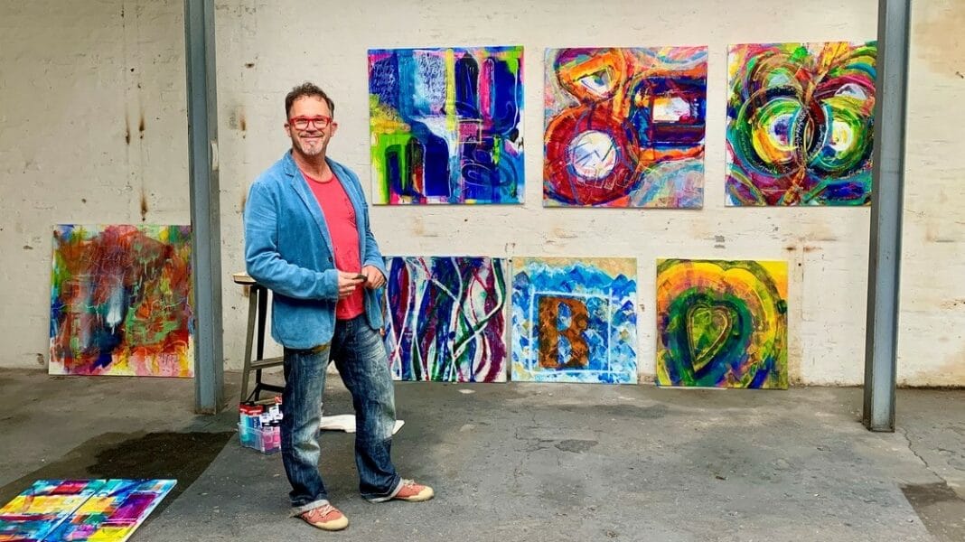 Daniel Kallauch steht vor einer weißen Wand, an der ein paar seiner Bilder hängen.