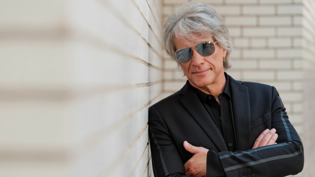 Jon Bon Jovi lehnt sich mit verschränkten Armen an eine Steinwand. Er trägt einen schwarzen Anzug und eine schwarze Sonnenbrille.