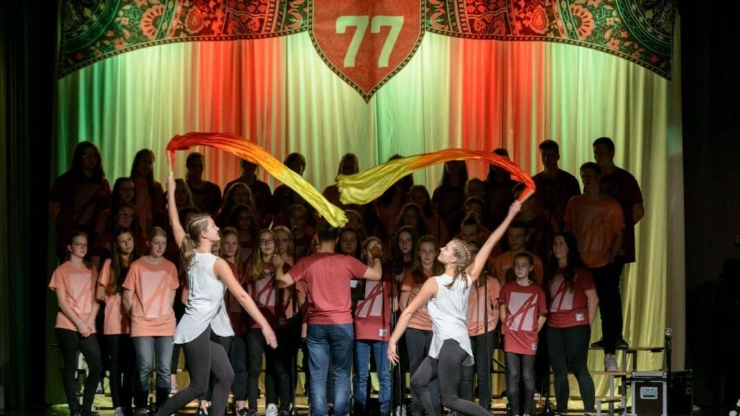 Zwei Mädchen schwingen bunte Bänder durch die Luft. Im Hintergrund singt ein Chor.