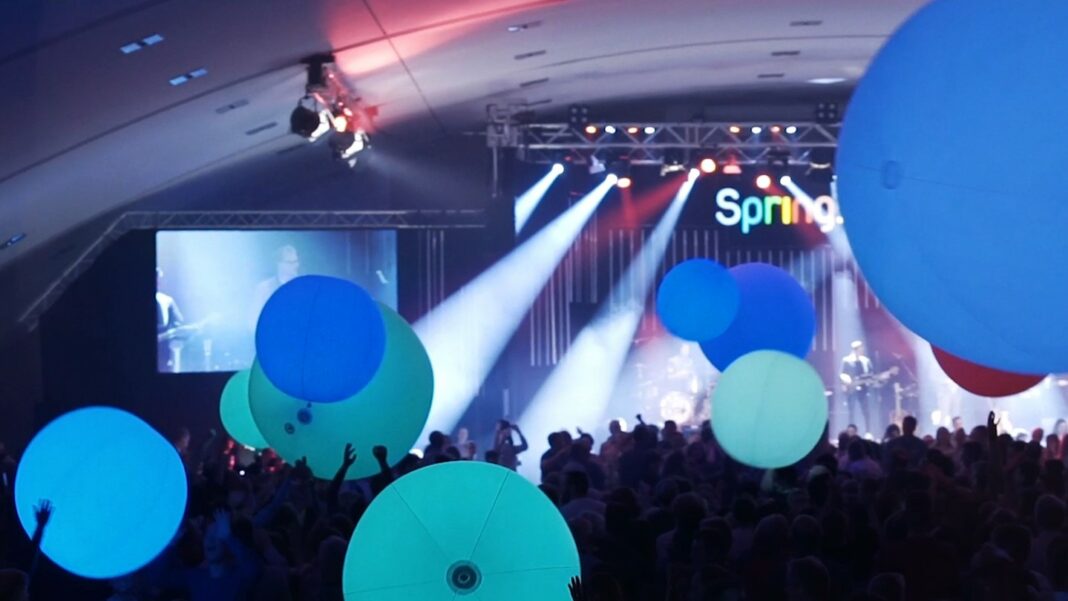Blaue leuchtende Luftballons schweben über einer Menschenmenge, die vor einer Bühne mit Lichtshow steht.