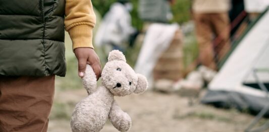 Ein Kind hält einen weißen Teddybären in seiner Hand. Im Hintergrund ist ein Zelt und Menschen zu sehen.