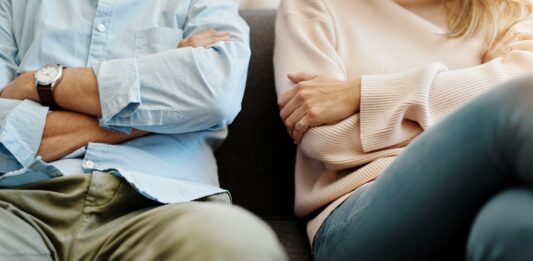 Ein Mann und eine Frau sitzen mit verschränkten Armen nebeneinander auf einem Sofa.