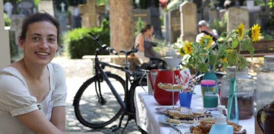 Eine Frau sitzt neben einem Tisch auf dem Kaffee und Kuchen steht. Im Hintergrund stehen Grabsteine.