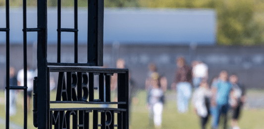 Blick auf das Eingangstor mit der Inschrift "Arbeit macht frei", im Hintergrund gehen Besucher über das Gelände des ehemaligen KZ Sachsenhausen.