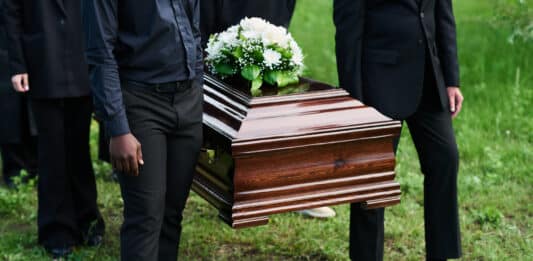 Männer in schwarzen Anzügen tragen einen Sarg mit Blumen drauf