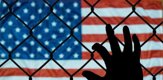 Eine Hand hält sich an einem Drahtzaun fest. Im Hintergrund ist die Flagge der USA zu sehen.