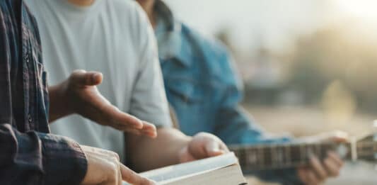 Mehrere Hände zeigen auf eine Bibel und gestikulieren. Im Hintergrund spielt jemand Gitarre.