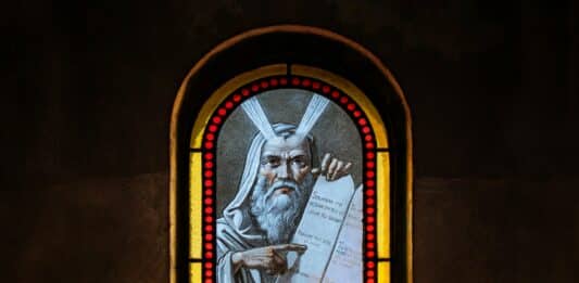 Auf einem Buntglasfenster ist ein alter Mann abgebildet. Er schaut ernst und zeigt mit dem Finger auf eine Tafel mit den Zehn Geboten.