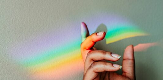 Eine Hand ist vor einer weißen Wand in die Luft gehoben. Darauf reflektiert sich ein Regenbogen.