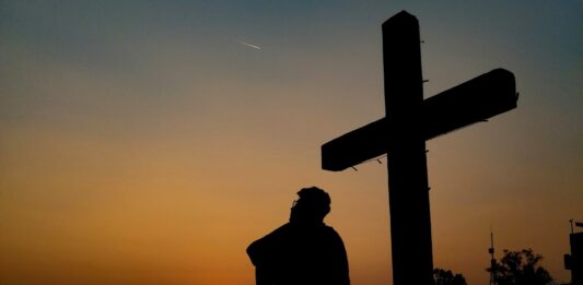 Die Silhouette eines Mannes, der vor einem Kreuz sitzt. Im Hintergrund ist ein Sonnenuntergang.