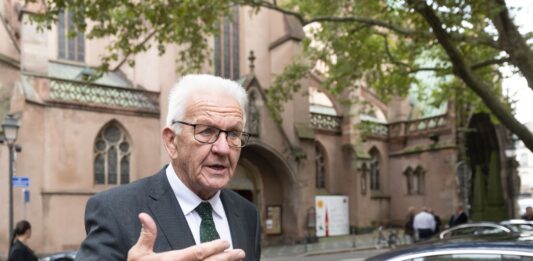 Winfried Kretschmann steht vor einer Kirche und erklärt etwas.