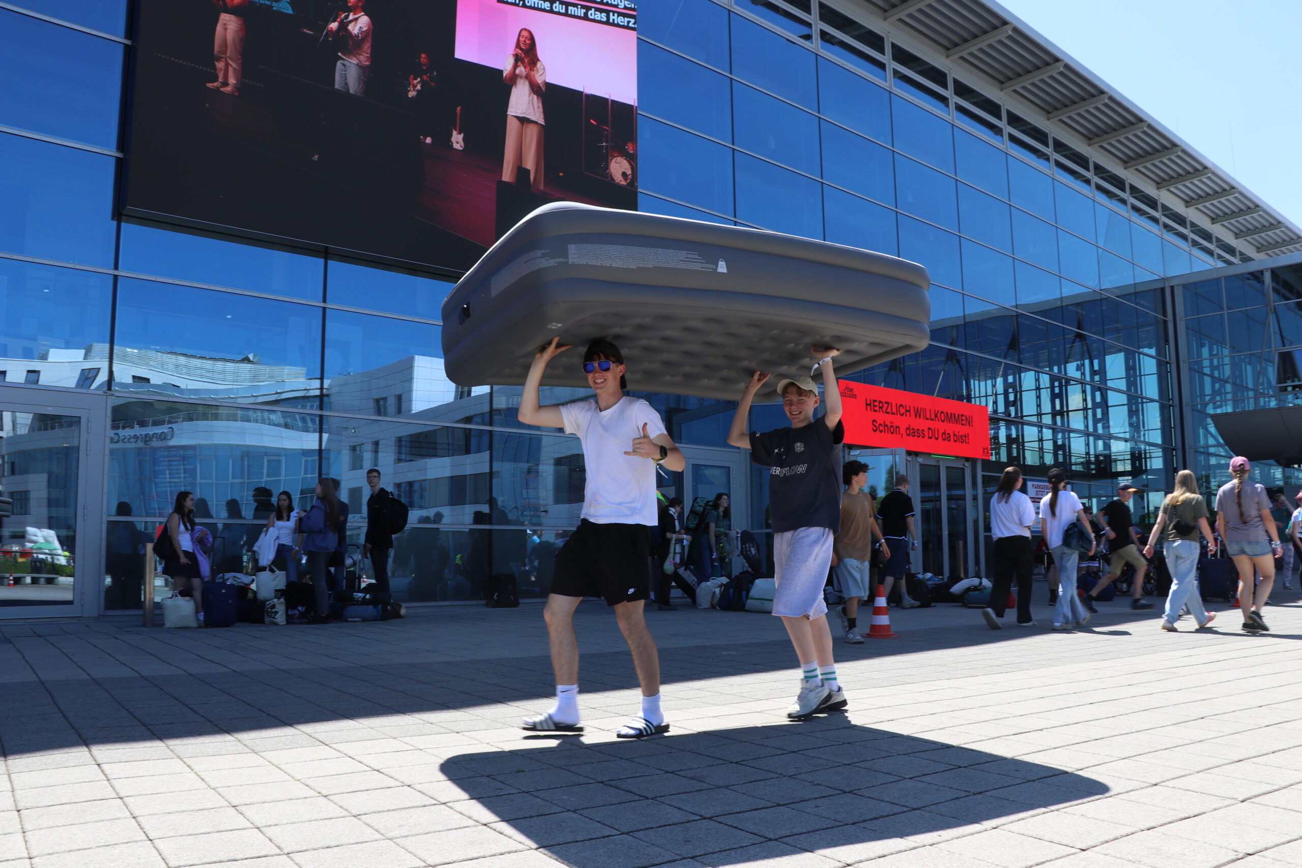 Zwei Jugendliche schützen sich mit einer Luftmatratze, die sie über ihrem Kopf tragen, vor der Sonne vor der großen Messe-Halle in Erfurt