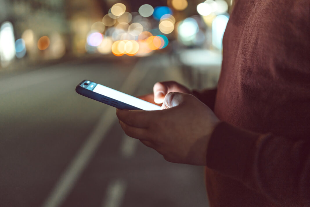 Eine Person tippt auf ihr Handy, verschwommene Straßenlichter sind im Hintergrund.