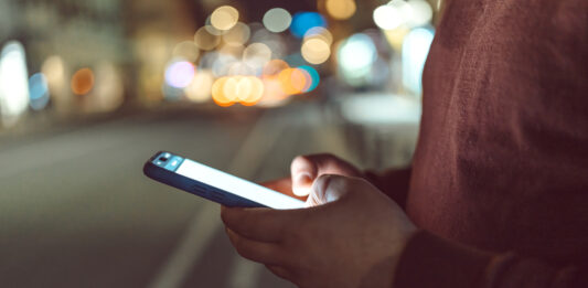 Eine Person tippt auf ihr Handy, verschwommene Straßenlichter sind im Hintergrund.